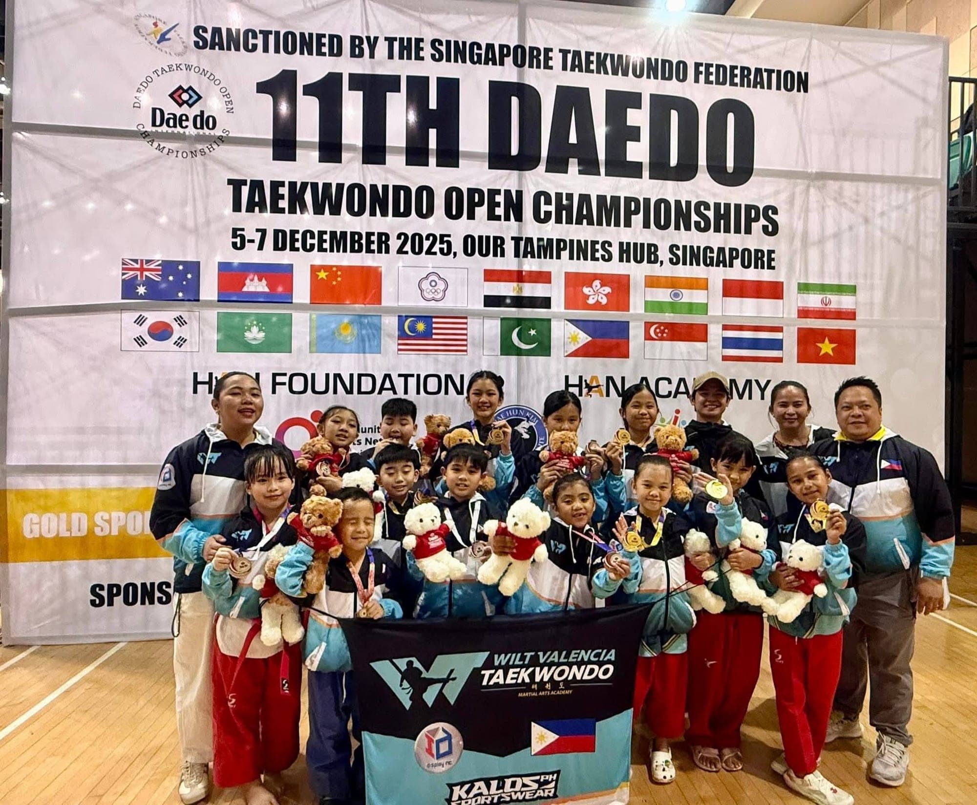 The WVT team with medals and teddy bear prizes in front of the 11th Daedo Taekwondo Open Championships banner in Singapore, holding the Wilt Valencia Taekwondo flag.