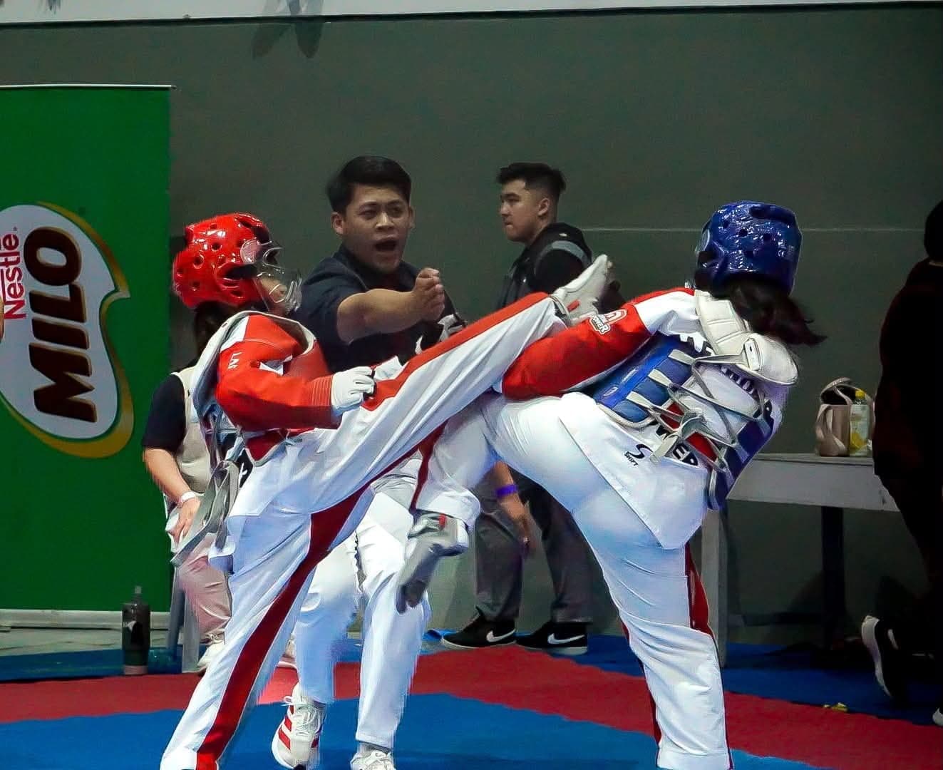 A WVT athlete in a red helmet lands a high roundhouse kick on her opponent during a sparring match, as the coach shouts instructions from behind.