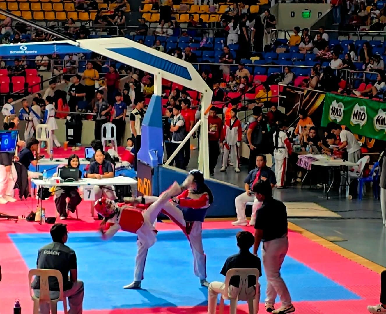 Two young athletes in headgear and chest protectors trading kicks on a tournament court while a WVT coach watches from the corner.