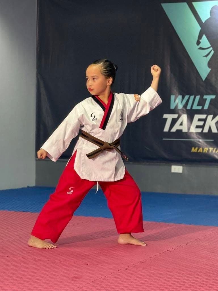 A young brown belt in a poomsae uniform holding a strong stance in front of the WVT banner.
